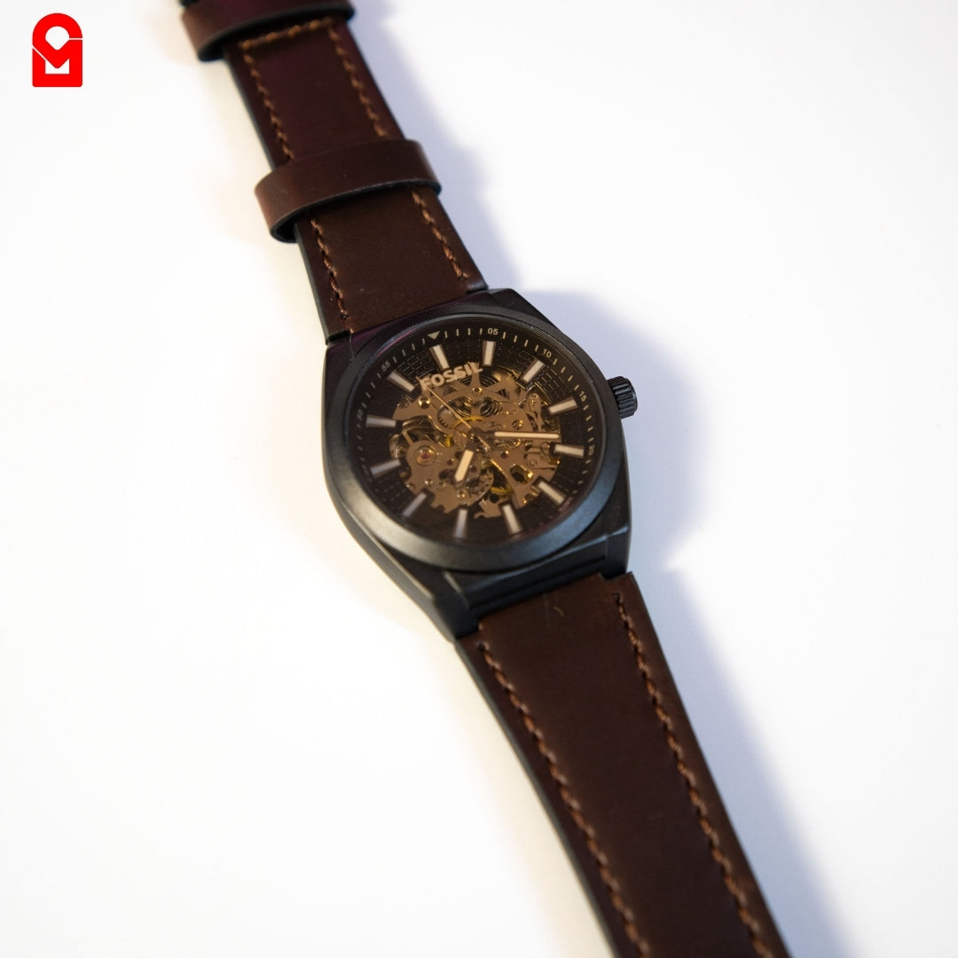 Black skeleton automatic watch with brown leather strap on white background.