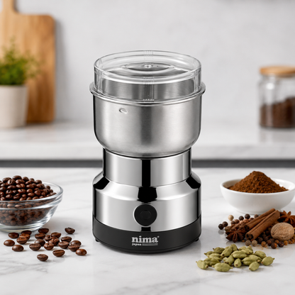 Compact electric coffee and spice grinder with stainless steel bowl displayed on a kitchen countertop alongside coffee beans, ground coffee, cardamom and whole spices.