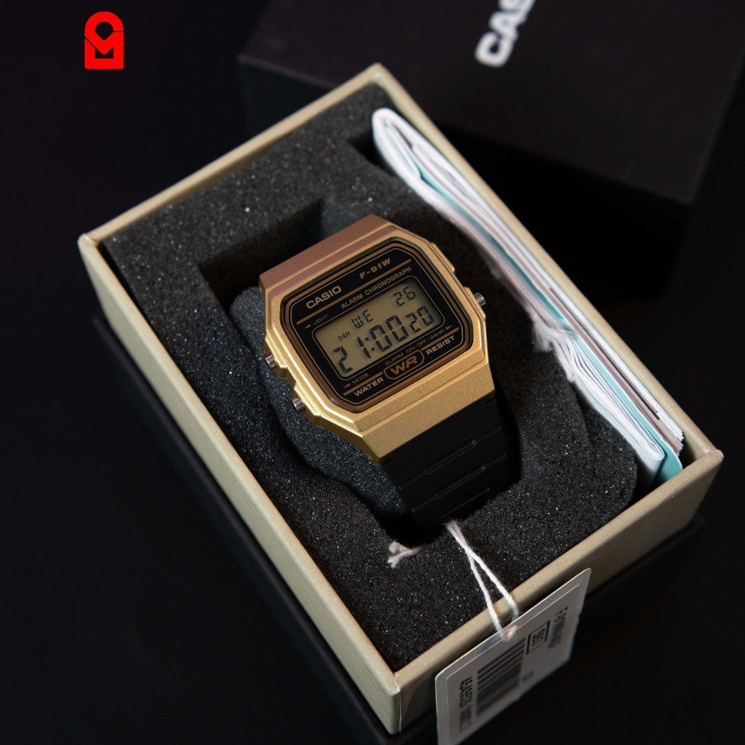 Gold tone digital rectangular watch with black resin strap displayed inside watch box.