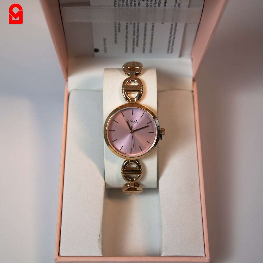 Analog women’s watch with pink dial and gold tone bracelet shown in packaging.