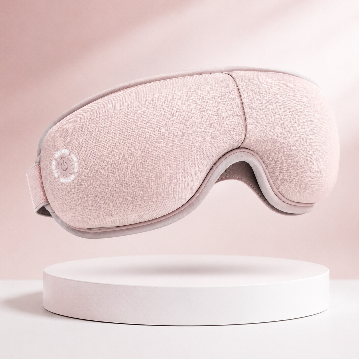 Soft pink eye massager mask with ergonomic contour design, adjustable strap, and rechargeable functionality, displayed on a clean white pedestal with a blush pink background for premium ecommerce presentation.