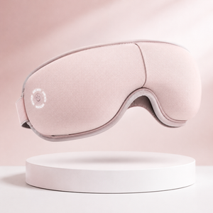 Soft pink eye massager mask with ergonomic contour design, adjustable strap, and rechargeable functionality, displayed on a clean white pedestal with a blush pink background for premium ecommerce presentation.