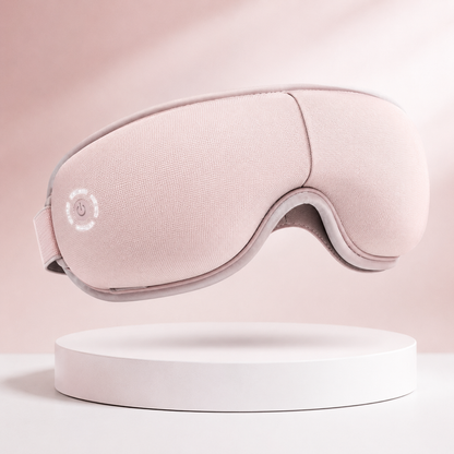 Soft pink eye massager mask with ergonomic contour design, adjustable strap, and rechargeable functionality, displayed on a clean white pedestal with a blush pink background for premium ecommerce presentation.
