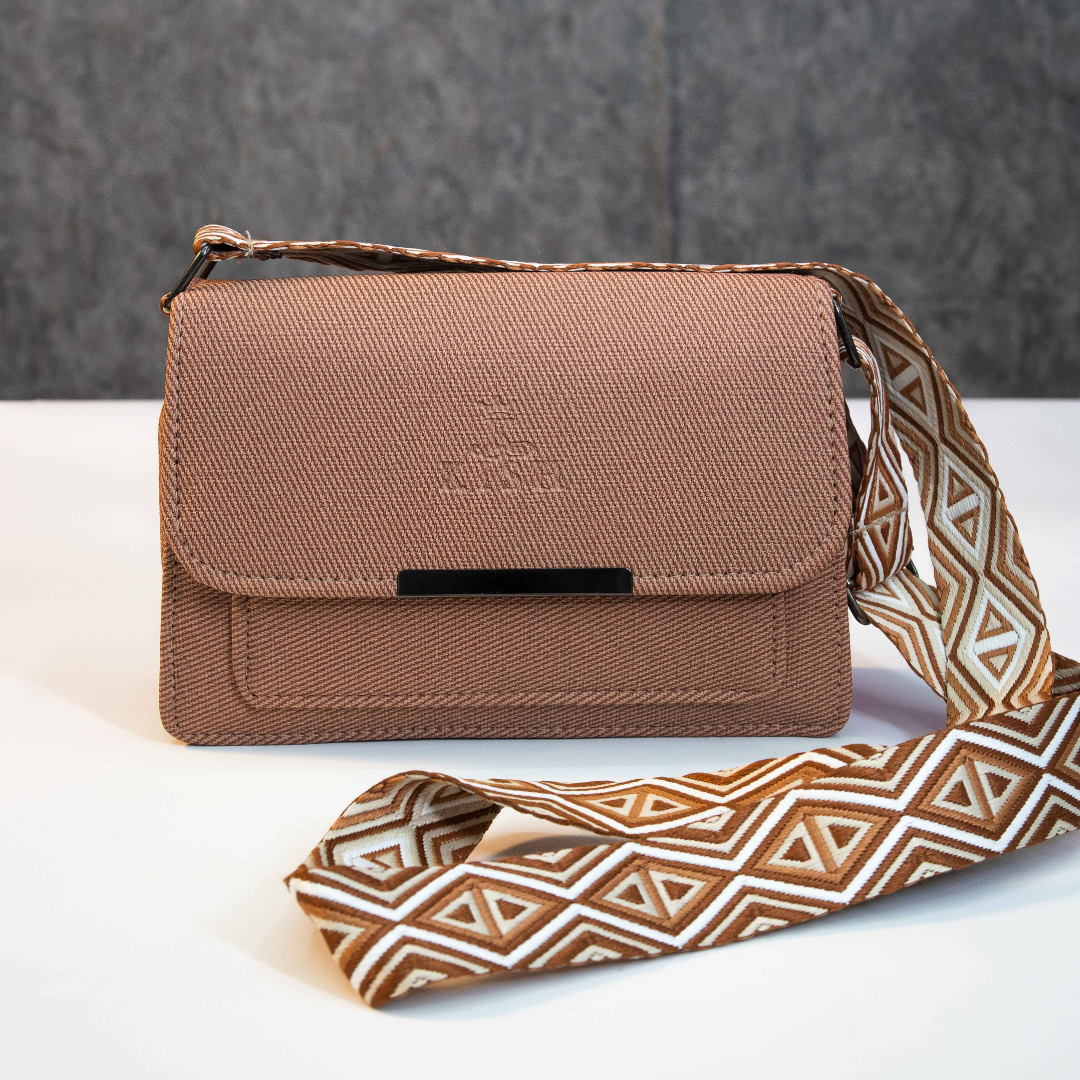Brown flap closure sling bag with wide patterned strap on flat surface.