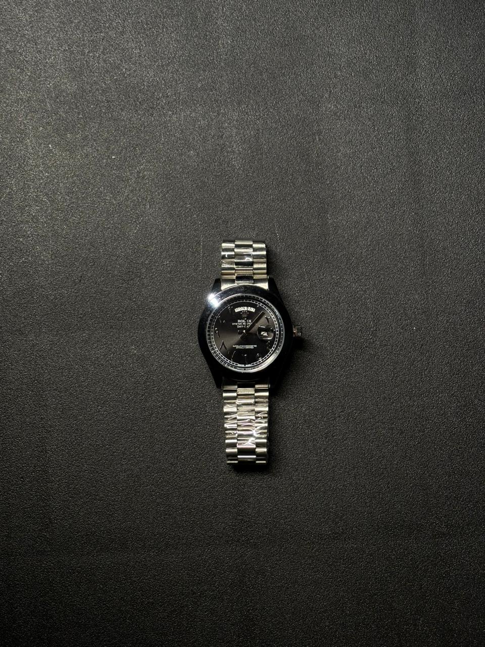Replica luxury day date watch with black dial, silver stainless steel bracelet, Arabic numerals and date magnifier displayed on dark textured surface