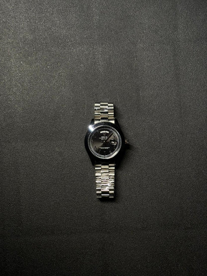 Replica luxury day date watch with black dial, silver stainless steel bracelet, Arabic numerals and date magnifier displayed on dark textured surface
