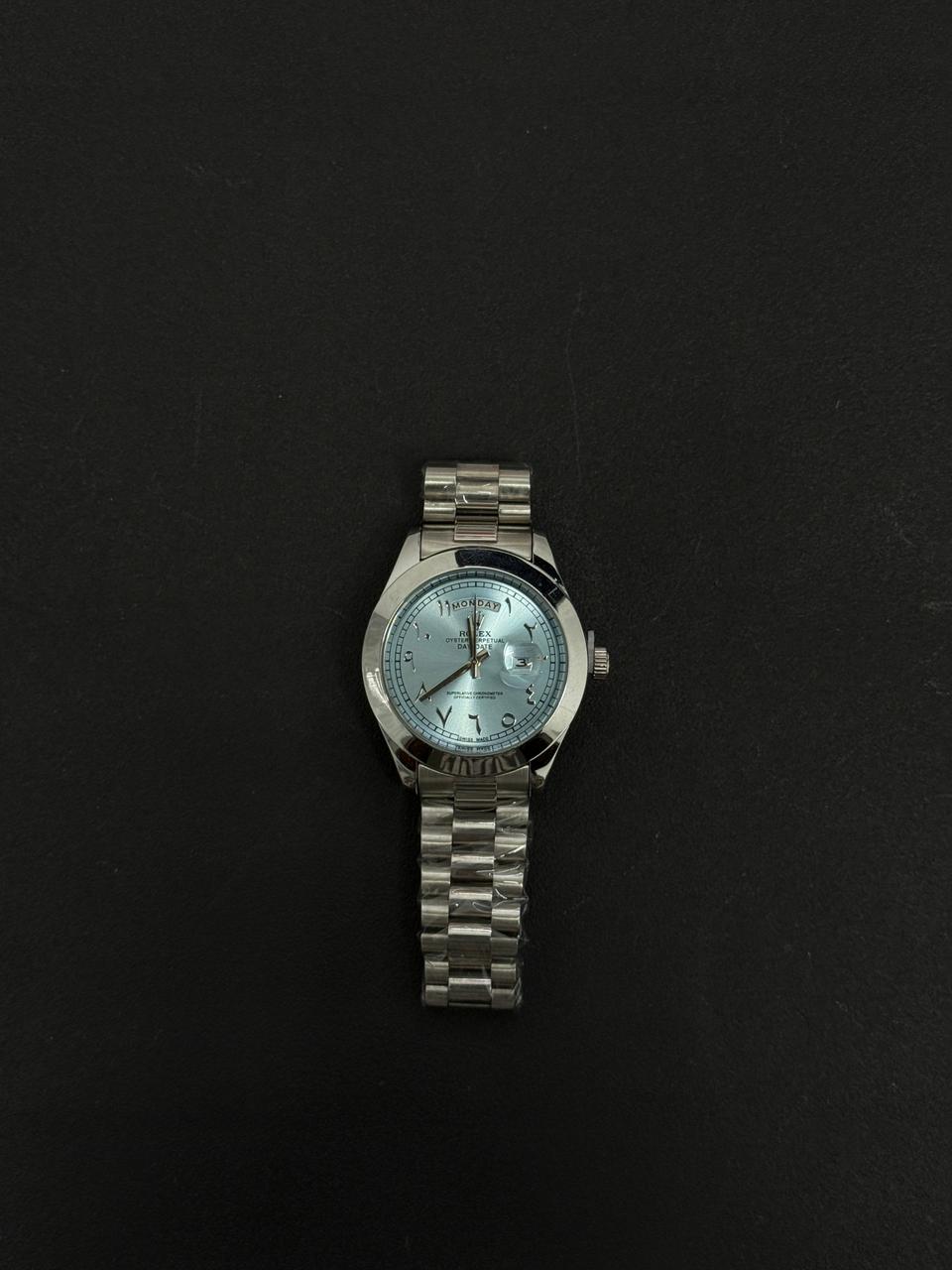 Replica stainless steel luxury watch with light blue dial, Arabic numeral markers, day date display and silver metal bracelet on black background