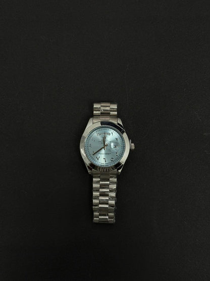 Replica stainless steel luxury watch with light blue dial, Arabic numeral markers, day date display and silver metal bracelet on black background