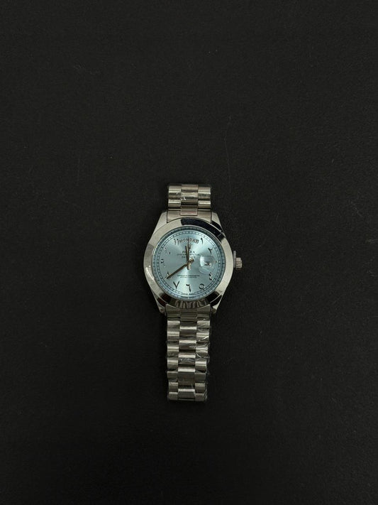 Replica stainless steel luxury watch with light blue dial, Arabic numeral markers, day date display and silver metal bracelet on black background