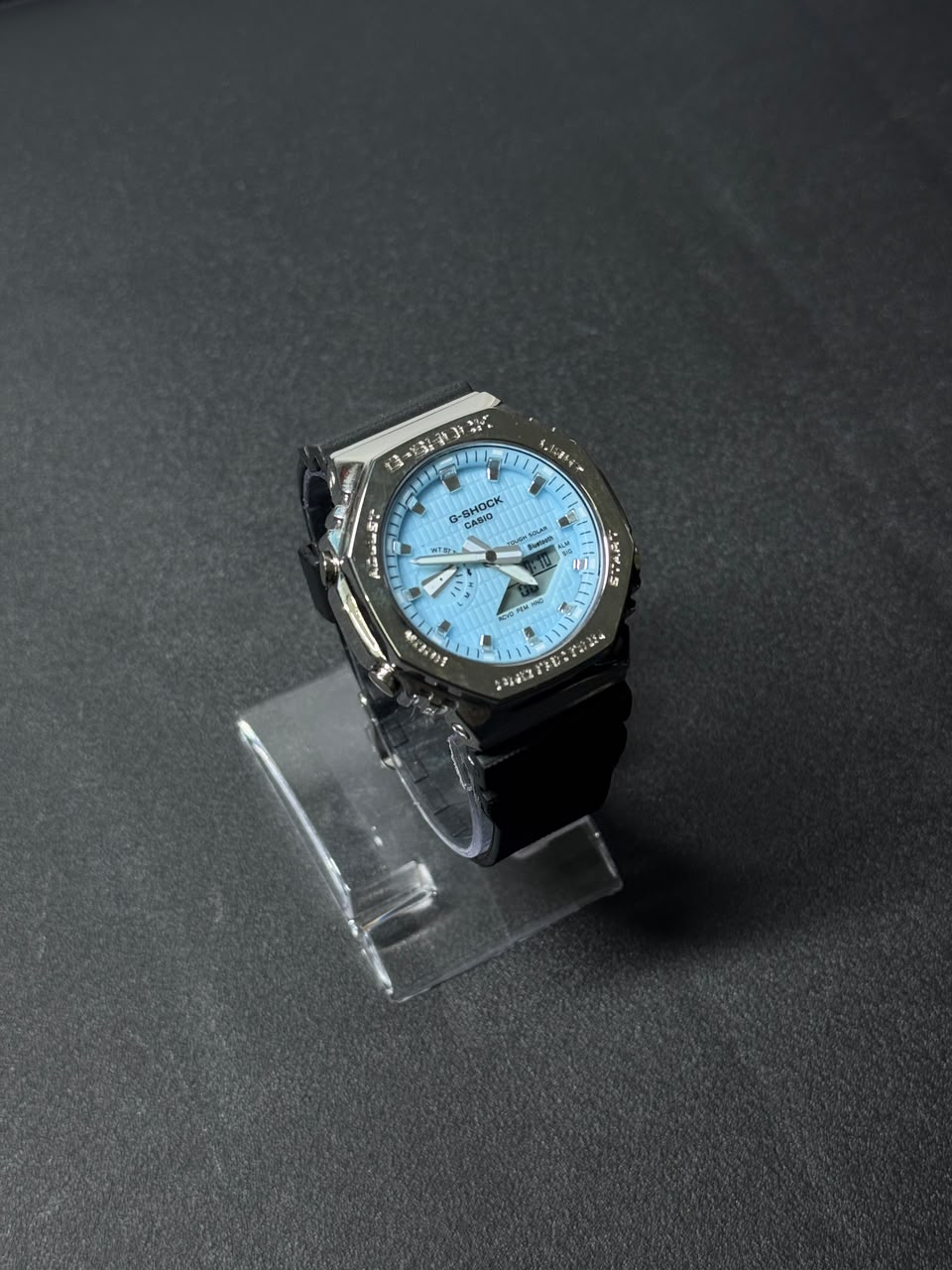 Replica watch with sky blue dial, octagonal bezel and black resin strap displayed on a clear stand against a dark premium background.