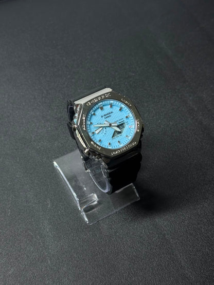 Replica watch with sky blue dial, octagonal bezel and black resin strap displayed on a clear stand against a dark premium background.