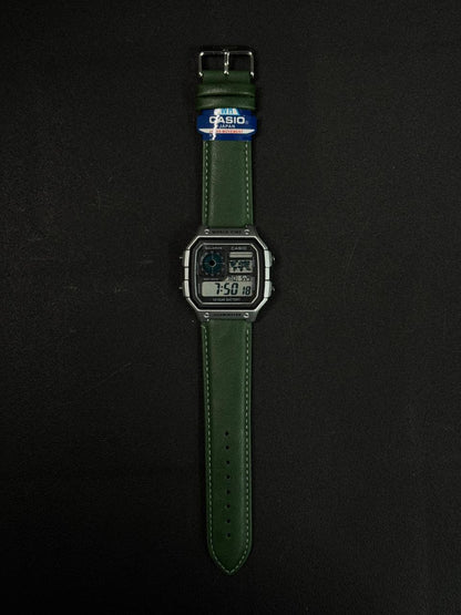 Replica digital watch with green leather strap and square black dial on dark background