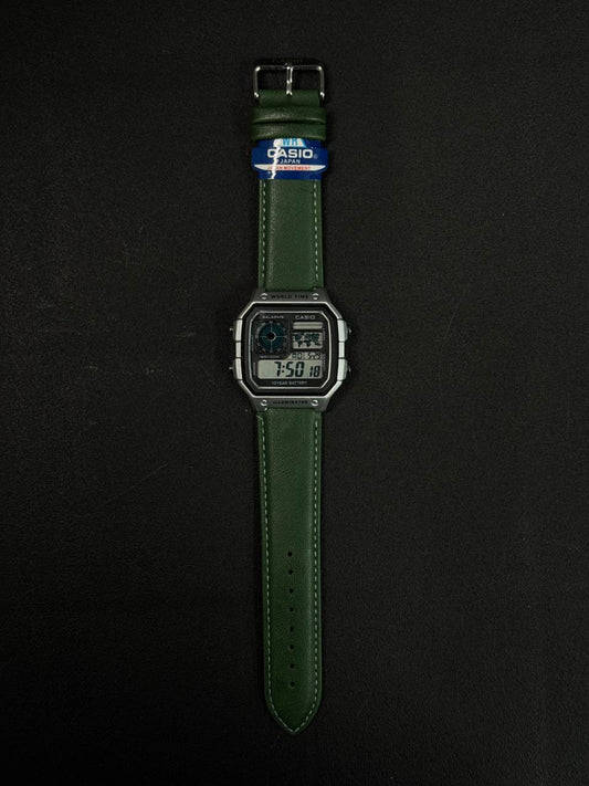 Replica digital watch with green leather strap and square black dial on dark background
