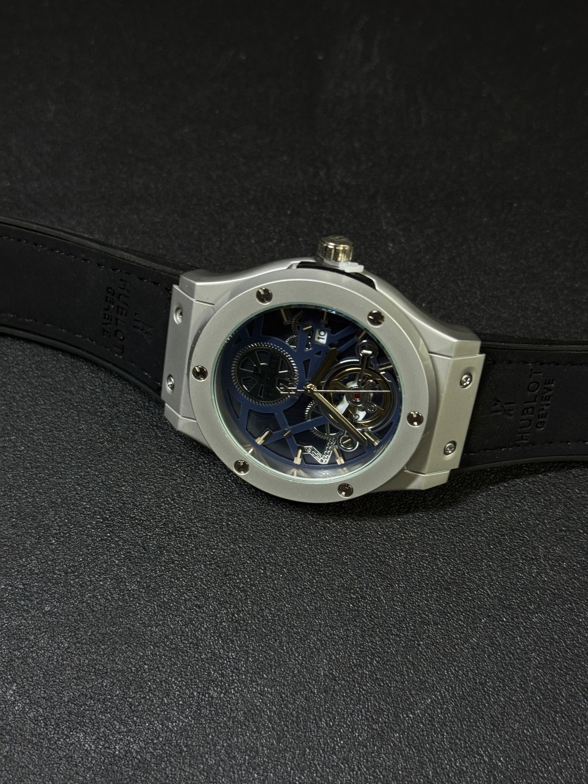 Luxury replica watch with blue skeleton dial, silver-tone case, black strap, and tourbillon-style open-heart display on dark background