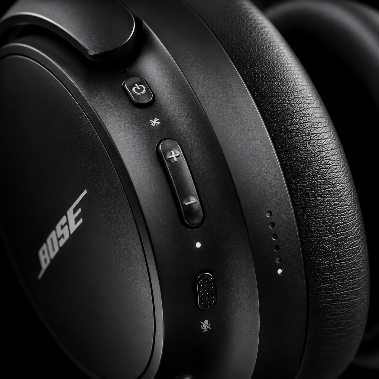 Close-up of premium wireless over-ear headphones showing onboard power button, Bluetooth pairing button, volume controls, microphone and USB-C charging port with soft cushioned ear pad and matte black finish.
