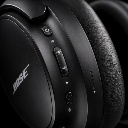 Close-up of premium wireless over-ear headphones showing onboard power button, Bluetooth pairing button, volume controls, microphone and USB-C charging port with soft cushioned ear pad and matte black finish.